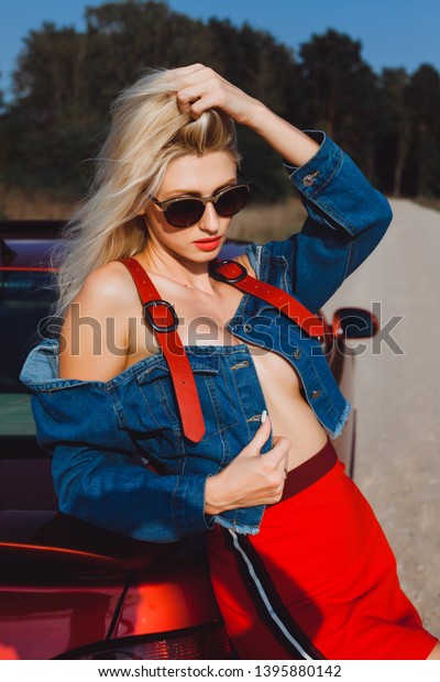 Glamor blonde with a good\
figure poses near a red car in a denim jacket and glasses in the\
summer