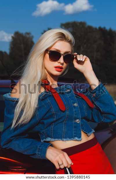 Glamor blonde with a good\
figure poses near a red car in a denim jacket and glasses in the\
summer