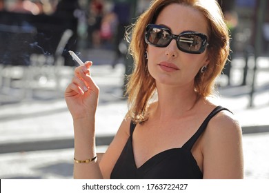 Glamor Caucasian Brunette Woman Wearing 50s Style Makeup Wearing A Black Dress And Sunglasses Having A Drink On A Terrace And Smoking A Cigarette Outside In The Center Of Brussels