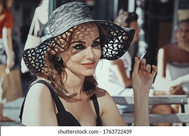 
Glamor Caucasian Brunette Woman Wearing 50s Style Makeup Wearing A Black Dress And A Sun Hat Having A Drink On A Terrace And Smoking A Cigarette Outside In The Center Of Brussels