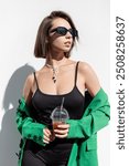 Glamorous beautiful fresh summer girl with bob haircut with vintage sunglasses in fashion green blazer with espresso tonic coffee near white wall outdoors in sunlight. Pretty lady on the street