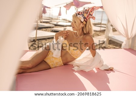 Glamorous caucasian young girl lies on beach bed with canopy on seashore. Beauty in yellow two-piece swimsuit and white shirt on top is resting on summer day. Rest time concept.