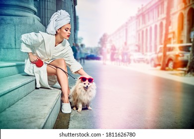 Glamorous Lady In A White Bathrobe With A White Towel On Her Head Sitting On The Steps Of A House With A Small Fluffy Dog On A Leash On The Street Of A Big City. Beauty, Fashion.