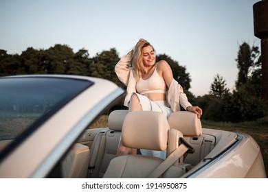 A glamorous luxury girl with blonde hair smiling while sitting on a white convertible. Young successful woman sitting in her white car