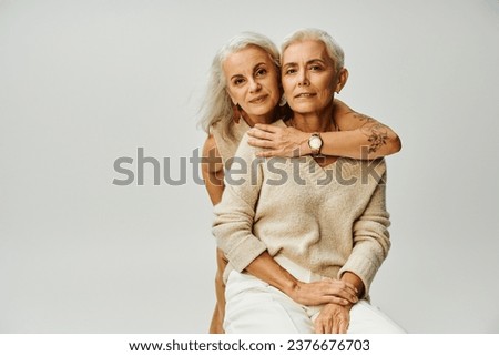 glamorous tattooed woman embracing smiling and stylish female friend sitting on grey, trendy seniors