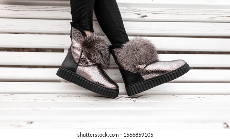 Glamorous winter stylish silver boots with gray fur. Modern young woman in black jeans and in fashionable shiny warm boots. New collection of beautiful trendy women's sneakers. Close-up.