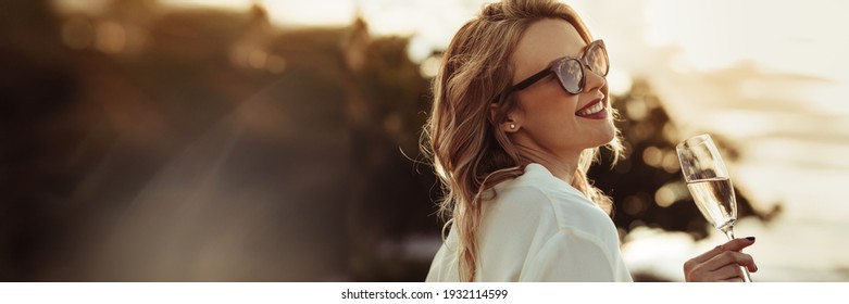 Glamorous woman having wine outdoors with large copyspace background. Beautiful female model wearing sunnglasses with a glass of wine.