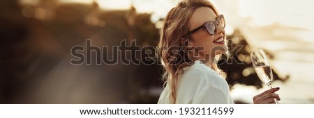 Glamorous woman having wine outdoors with large copyspace background. Beautiful female model wearing sunnglasses with a glass of wine.