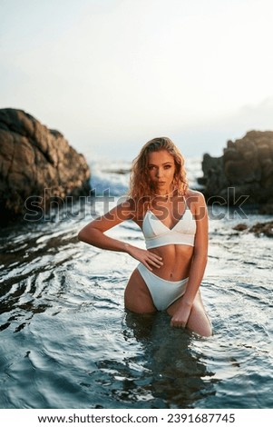 Glamorous woman poses in chic white bikini on sea shore, sunlit waves caress her sun-kissed skin, exemplifying seaside fashion elegance.
