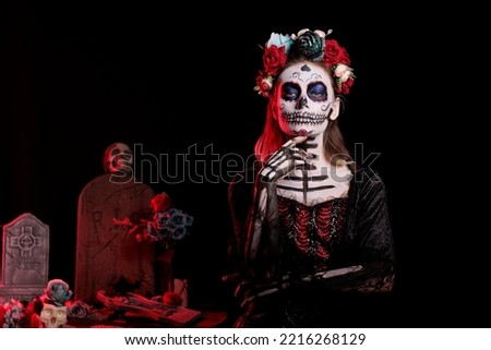 Glamorous woman wearing traditional body art on halloween day of the dead, having flowers crown. Female model with black and white make up and holy santa muerte costume on holiday.