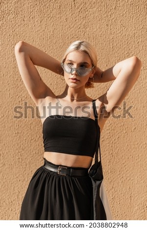 Glamorous young beautiful hipster girl with sunglasses and short hair in a summer stylish black top and skirt with a bag stands near a beige wall on the street in sunny day