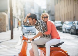 Glamorous  Young Couple Riding  A Vintage Scooter In The Street, Man Wears A Hat And Woman Has A Topknot And Sunglasses