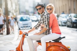 Glamorous  Young Couple Riding  A Vintage Scooter In The Street, Man Wears A Hat And Woman Has A Topknot And Sunglasses