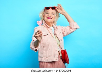 glamour awesome blonde senior lady taking off sunglasses, while eating lollipop. close up portrait. isolated blue background. beauty , happiness