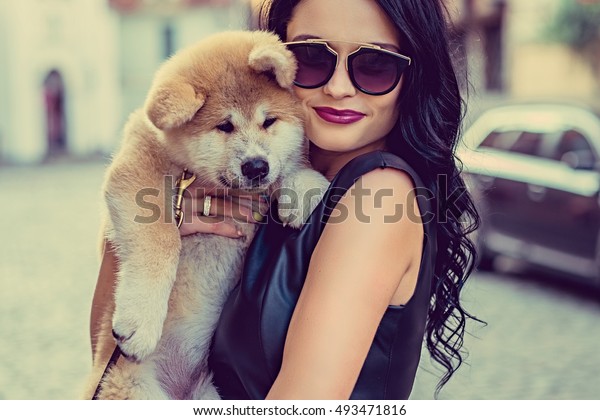 Glamour brunette female in sunglasses holding a\
dog puppy on a street in a\
town.\
