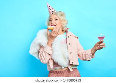 Glamour Old Lady In Party Hat Holding A Glass Of Wine And Eating Yummy Cake. Isolated Blue Background. Studio Shot. People, Happiness