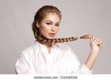 Glamourous young blond caucasian girl with healthy shiny long hairs in pigtail hold braid in hand. Alluring female with stylish plait posing on gray studio background. Haircare treatment cosmetics ad
