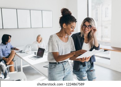 Gland Blonde Secretary In Glasses Looks At African Female Manager Which Holding Report. Indoor Portrait Of Stylish Black Woman In Jeans Spending Time In Office With Asian And European Colleagues.