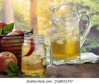 Glass of apple juice with apple slice and ice and a basket of apples and pitcher on a wooden table with rural summer background