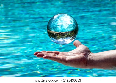 Glass Ball Hovers Over A Hand In Front Of Water. Background With Mirrored Cloudy Sky And Trees