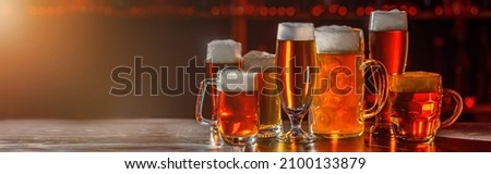Glass of beer on the bar counter. Jugs, mugs, pints of brew beverage, ale, cider on the wooden table in pub, bar. Backlit dark room, showcase with craft beer bottles in the vintage brewery.