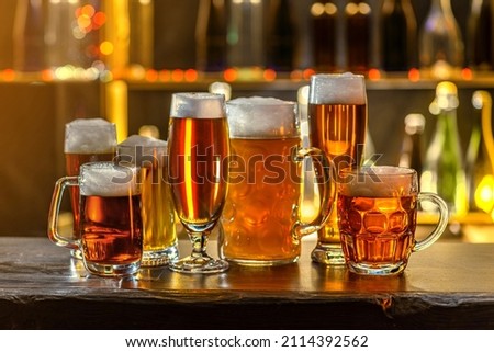 Glass of beer on the bar counter. Jugs, mugs, pints of brew beverage, ale, cider on the wooden table in pub, bar. Backlit dark room, showcase with craft beer bottles in the vintage brewery.
