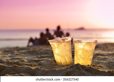 Glass Of Beer On Beach Sand At Sunset With Cloud Of People