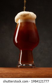 Glass Of Beer On Dark Background. Dark Ale Beer With Overflowing Foam Head. Stream Of Stout Beer Pours Into A Beer Glass On A Bar Counter.	