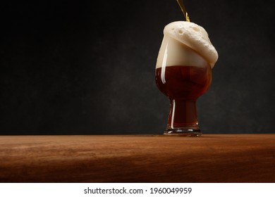 Glass Of Beer On Dark Gray Background. Dark Ale Beer With Overflowing Foam Head. Stream Of Stout Beer Pours Into A Beer Glass On A Bar Counter.