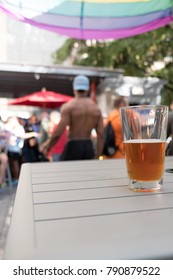 Glass Of Beer On A White Table Outside On A Rooftop Bar In NYC. Gay Men At A Gay Rooftop Bar In New York City On A  Weekend Afternoon For Day Drinking On Roof Top. Shirtless Black Waiter In Background