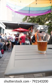 Glass Of Beer On A White Table Outside On A Rooftop Bar In NYC. People At A Gay Friendly Rooftop Bar In New York City On A Sunny Weekend Afternoon For Day Drinking On Roof Top.