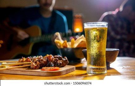 The glass of beer put on wooden table, there are musician on background, focus at glass of beer, shallow depth of field.
