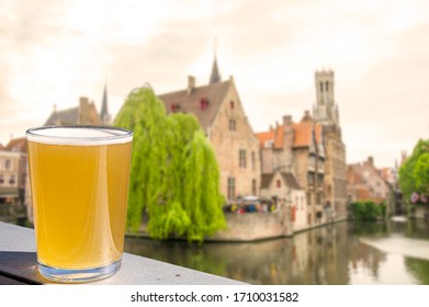 Glass of beer with view of historic houses, canal and belfry tower in city center in Bruges, Belgium. 