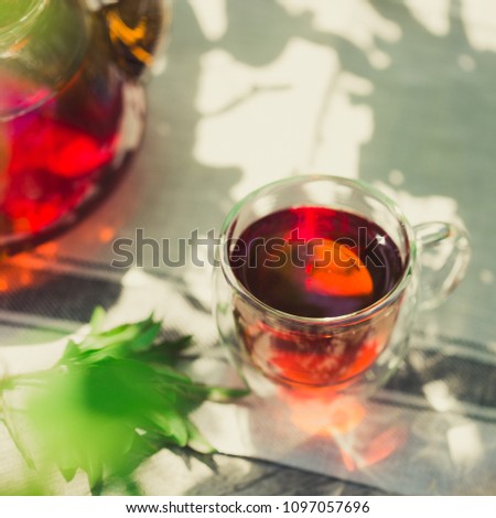 Glass of black tea in double bottom glass under the shade of the tree with green leaves under sun light, summer hot drink with glass kettle of strong tea on wooden table and tea cloth, copy space