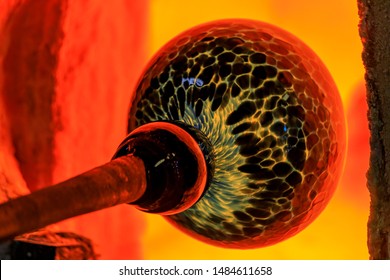 Glass blower manufacturing process, working on a bubble of melted glass on a rod by heating it up by hand in a kiln at a glass maker's workshop