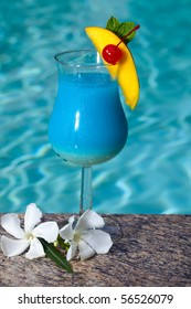 Glass Of Blue Hawaiian Cocktail On Swimming Pool Side Garnished With Mango Wedge And Maraschino Cherry