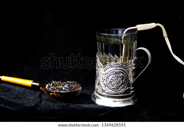 In a glass of boil water advisory is a vintage\
heater. Near painted spoon with flower tea. The image on a black\
background, illuminated by the sun. Concept - breakfast with tea\
drinking while travelin