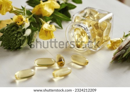 A glass bottle with capsules of evening primrose oil spilled on a table, with blooming Oenothera biennis plant in the background