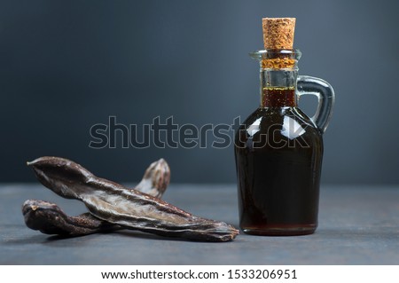 glass bottle of carob molasses and carob pods on rustic background, locust bean healthy food, Ceratonia siliqua ( harnup )