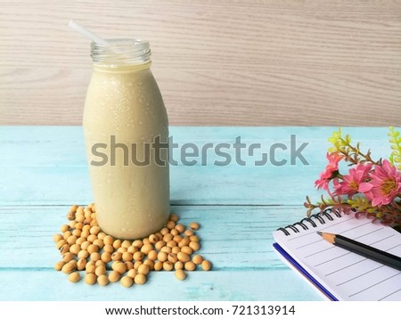 A glass bottle of delicious cold soy milk have are soybeans seed around base ,near a notebook ,a pencil and a bouquet of pink flowers on a blue wood table