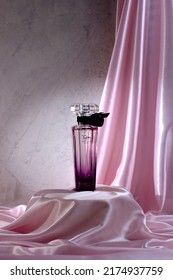 A Glass Bottle Of Midnight Rose Perfume By Lancome Stands On A Podium Covered With A Pink Silk Cloth Against A Stone Slab Background