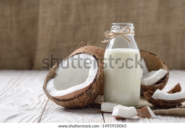 Glass bottle of milk or yogurt on hemp\
napkin on white wooden table with coconut\
aside