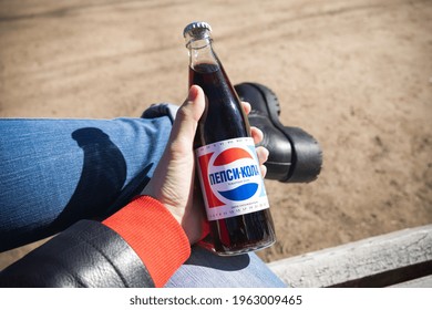 Glass bottle of Pepsi with inscription in Russian in hands of person sitting on bench with leg in blue rolled up jeans and black boots - Russia, Saint Petersburg, April 2021