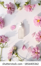 Glass Bottle With Pipette Next To Fresh Flowers And Petals On White Background. Natural Beauty Product. Flat Lay, Top View.