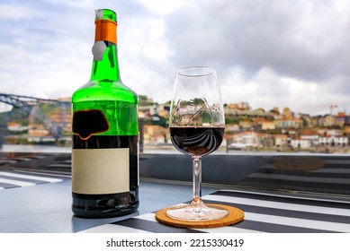 Glass And Bottle Of Red Ruby Port Wine At An Outdoor Restaurant In Porto, Portugal The Douro River And Dom Luis I Bridge Blurred In The Background