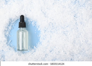 Glass Bottle With Serum, Essential Oil On Blue Background On The Snow. Gentle Moisturizing Skincare Product The Cold Winter Season. Cosmetic For Face And Hand Skin Care With Vitamins. Copy Space