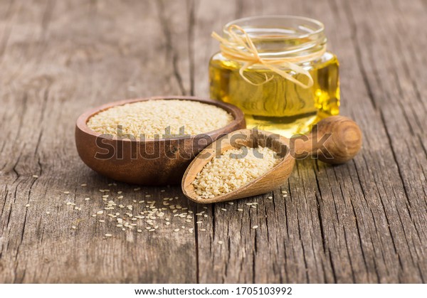 Glass bottle of sesame oil and raw sesame\
seeds in wooden shovel with burlap sack on wooden table. Uncooked\
sesame background concept with copy\
space