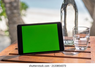 Glass Bottle And A Glass Of Water On The Background Of The Beach In The Maldives. Tablet With Green Screen. Chromakey, Mockup. 