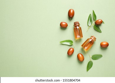 Glass bottles with jojoba oil and seeds on green background, flat lay. Space for text