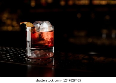 Glass of a Boulevardier cocktail with big ice cube and orange zest on the steel bar counter on the dark blurred background